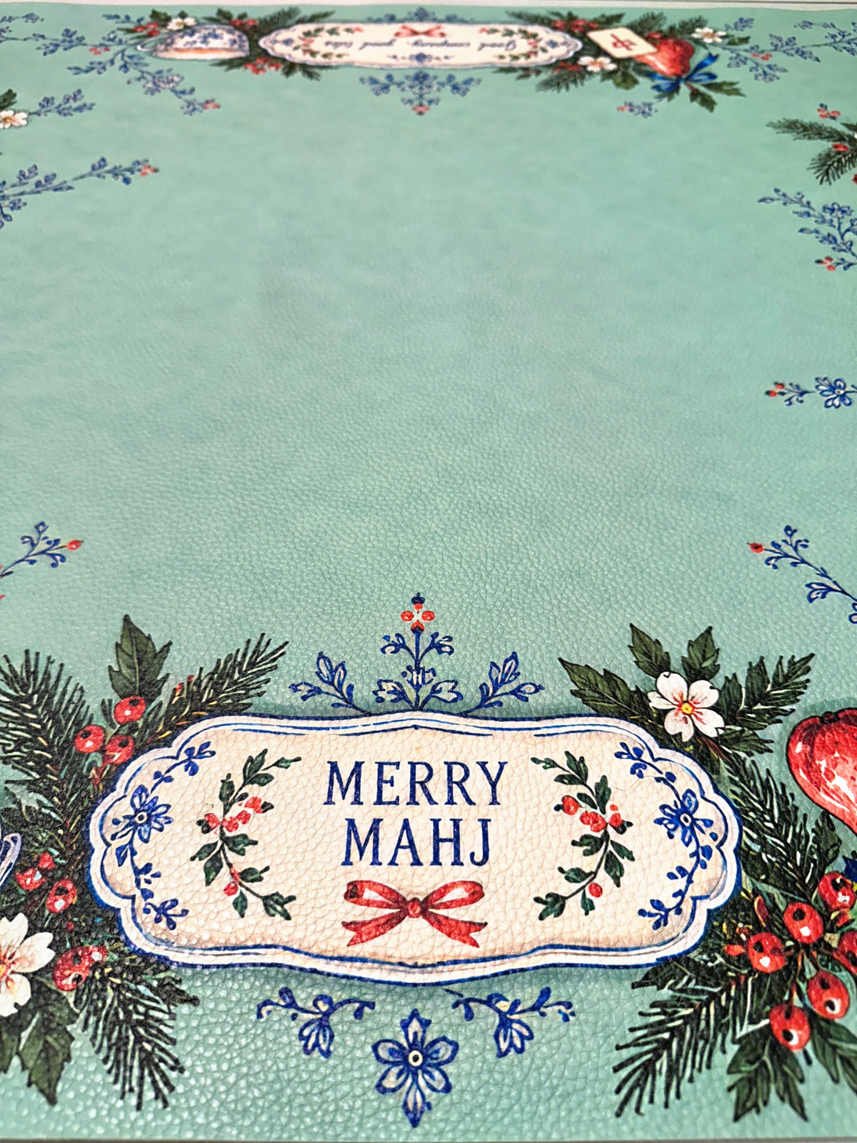 Close-up of Merry Mahj lettering with blue floral flourishes and Christmas greenery on a green Mahjong mat