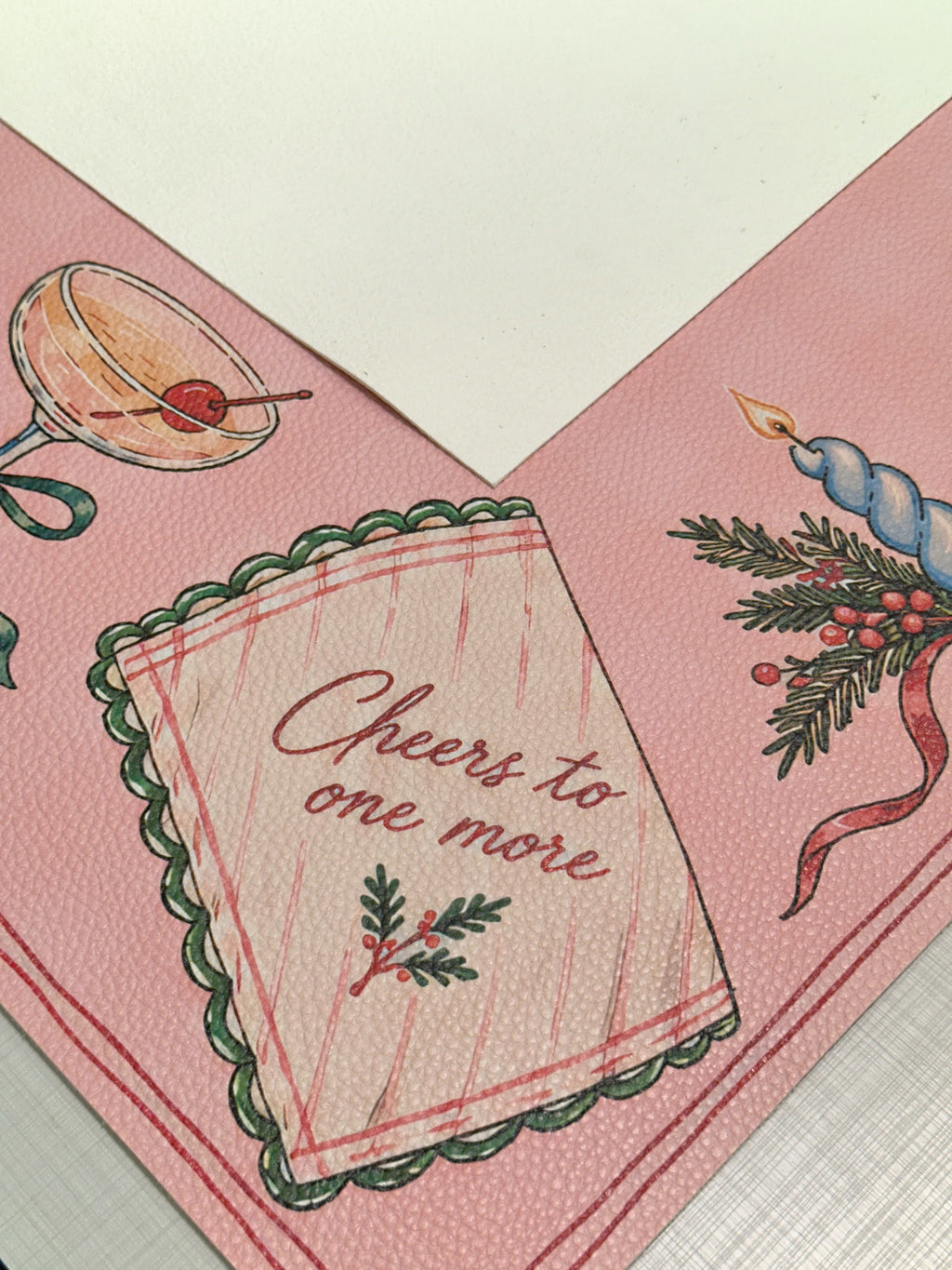 Cream suede backing beside the pink Merry Mahj mat with a Cheers to one more napkin and cocktail illustration