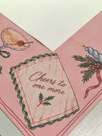 Cream suede backing beside the pink Merry Mahj mat with a Cheers to one more napkin and cocktail illustration