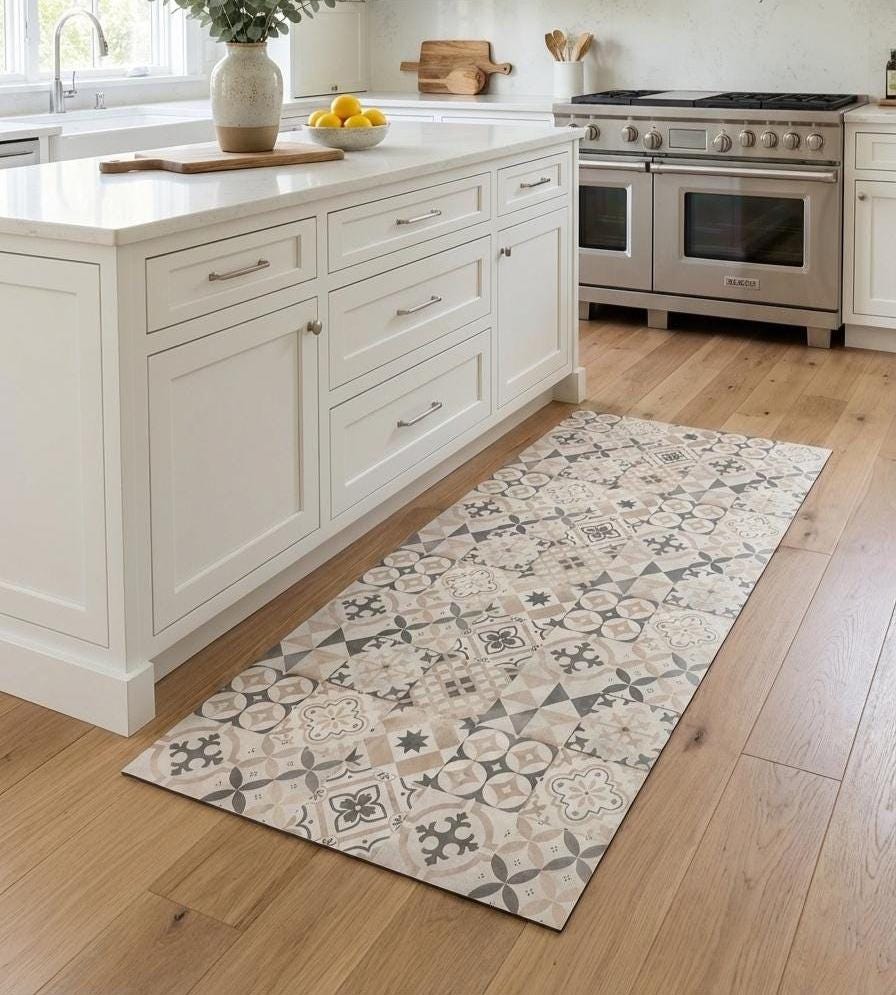 May include: A rectangular kitchen floor mat with a geometric pattern in shades of beige, gray, and taupe. The mat is placed in front of a white kitchen island with a wooden floor. The pattern includes floral and geometric shapes.